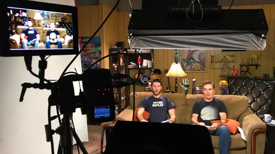 Talk-show style set with hosts seated on a couch under studio lights and cameras